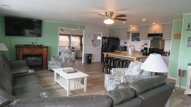 A living room with a sofa, a coffee table, and a TV. There is a kitchen area with a stove, refrigerator, and sink. - LastMinuteSpecial/OceanFront/Pool/HotTub/XBOXoneS/ping pong table (Nags Head)