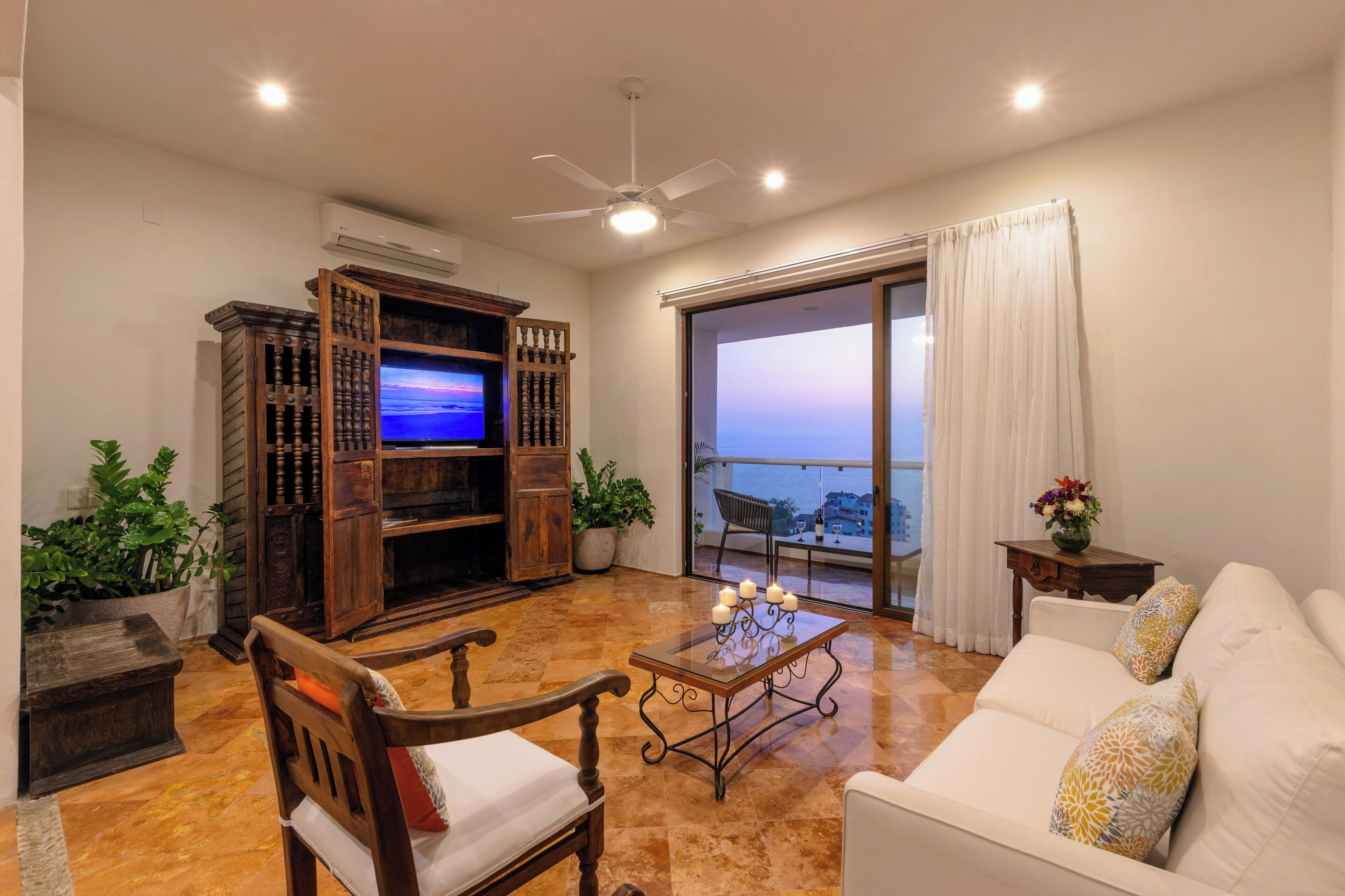 A living room with a TV, wooden furniture, a white sofa, and a balcony with a view.