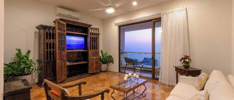 A living room with a TV, wooden furniture, a white sofa, and a balcony with a view.