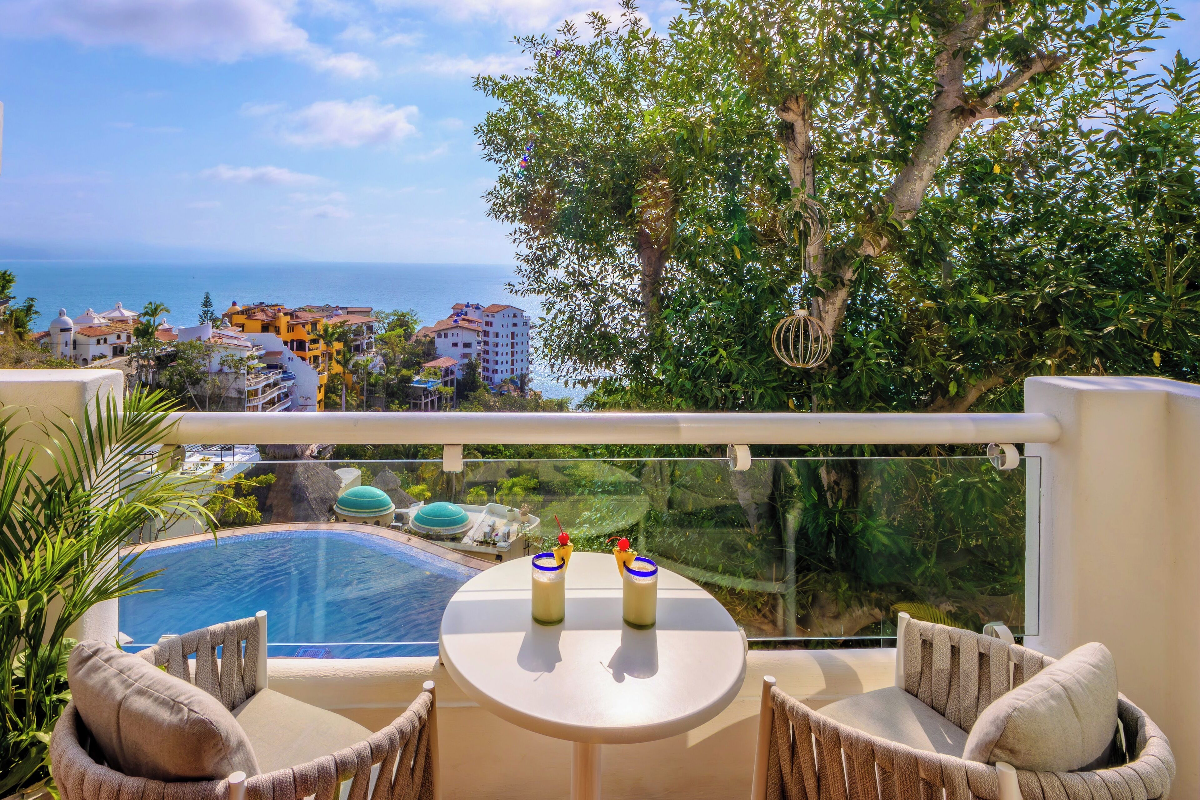 A balcony with a pool, two drinks on a table, and a view of the ocean and buildings.