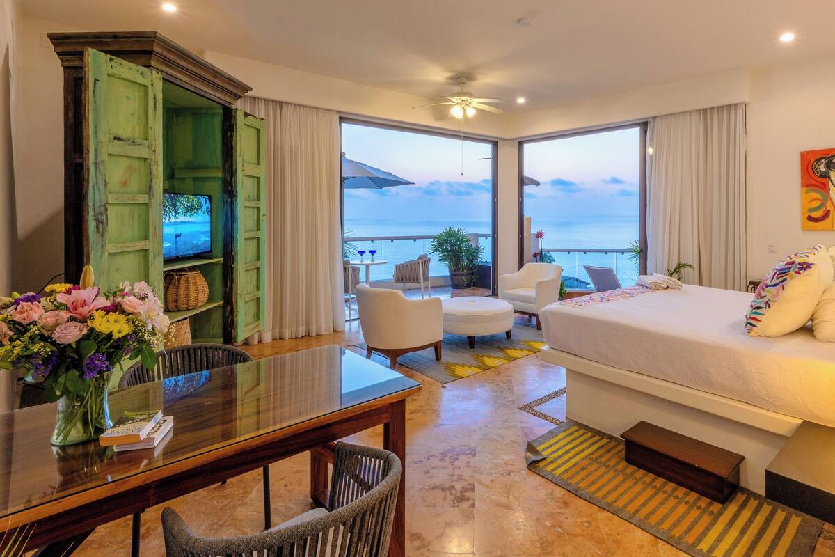 Ein Hotelzimmer mit Bett, Schreibtisch, Stuhl, einem Balkon mit Meerblick und einem Bild an der Wand.