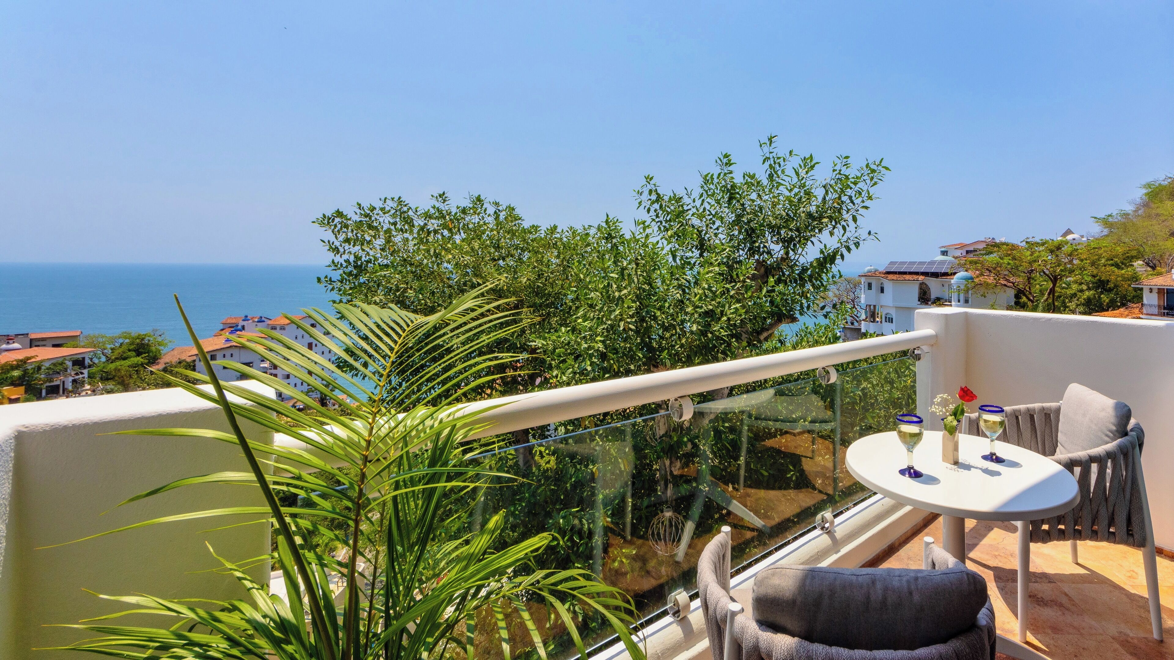 A balcony with a glass railing overlooking a coastal view, a table with a glass of wine, and a chair.