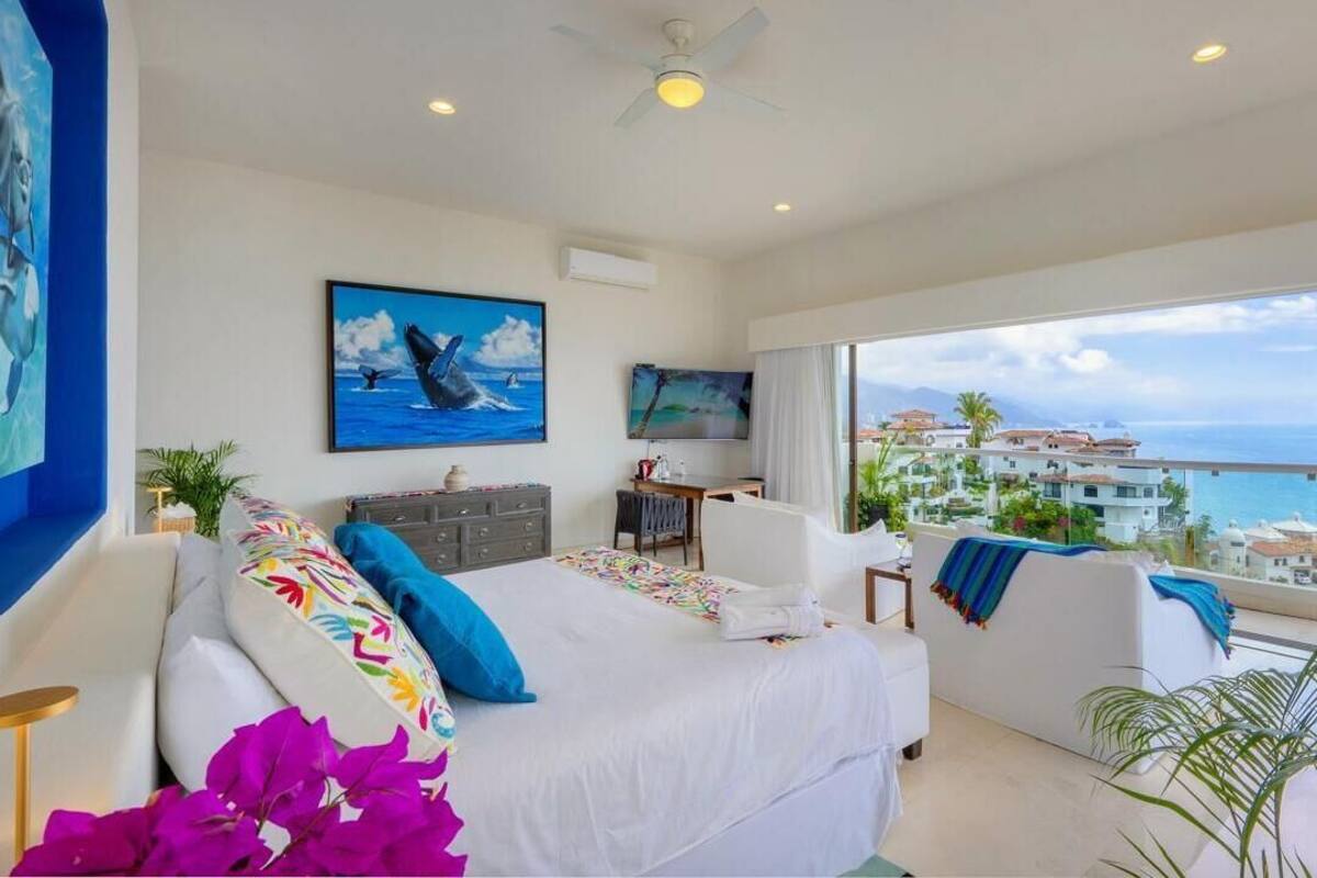 Ein Schlafzimmer mit einem großen Bett, einem Schreibtisch und einem Fernseher. Es bietet Blick auf das Meer und Gebäude.