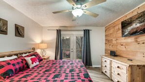 A bedroom with a bed, a ceiling fan, a nightstand, a lamp, a dresser, and a window with curtains.