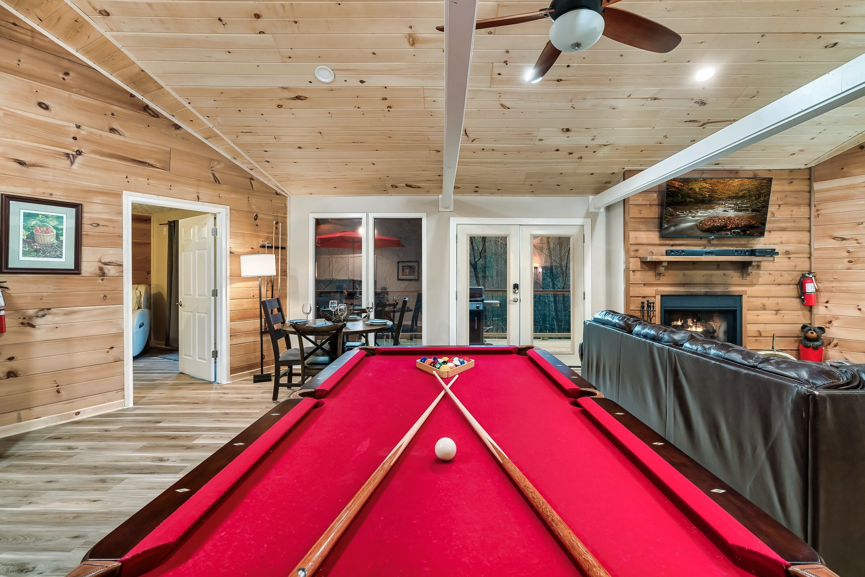 A pool table in a wooden room with a ceiling fan, a fireplace, and a dining area visible through glass doors.