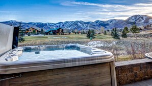 Un jacuzzi con recinto de madera, pared de piedra y vista a una cadena montañosa con casas al fondo.