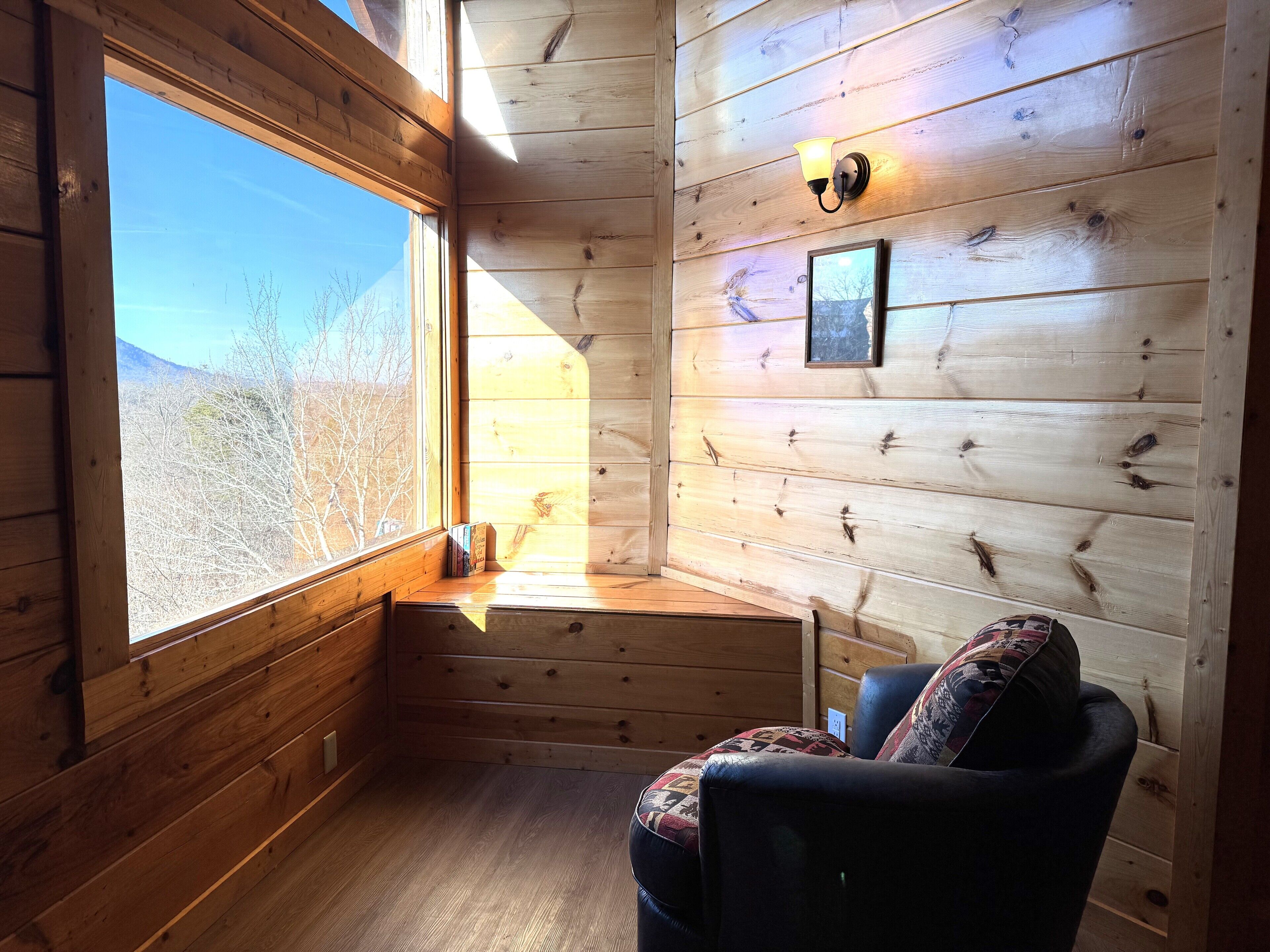 A cozy cabin room with a large window, wooden walls, a cushioned chair, and a wall-mounted light fixture.