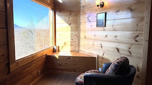 A cozy cabin room with a large window, wooden walls, a cushioned chair, and a wall-mounted light fixture. - Nature's View, Beautiful 3-Story Cabin; 3 Bdrms w/King Beds; Free WiFi (Sevierville)