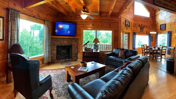 A cozy cabin interior with a stone fireplace, leather seating, a flat-screen TV, and large windows offering a view of the outdoors. - Nature's View, Beautiful 3-Story Cabin; 3 Bdrms w/King Beds; Free WiFi (Sevierville)