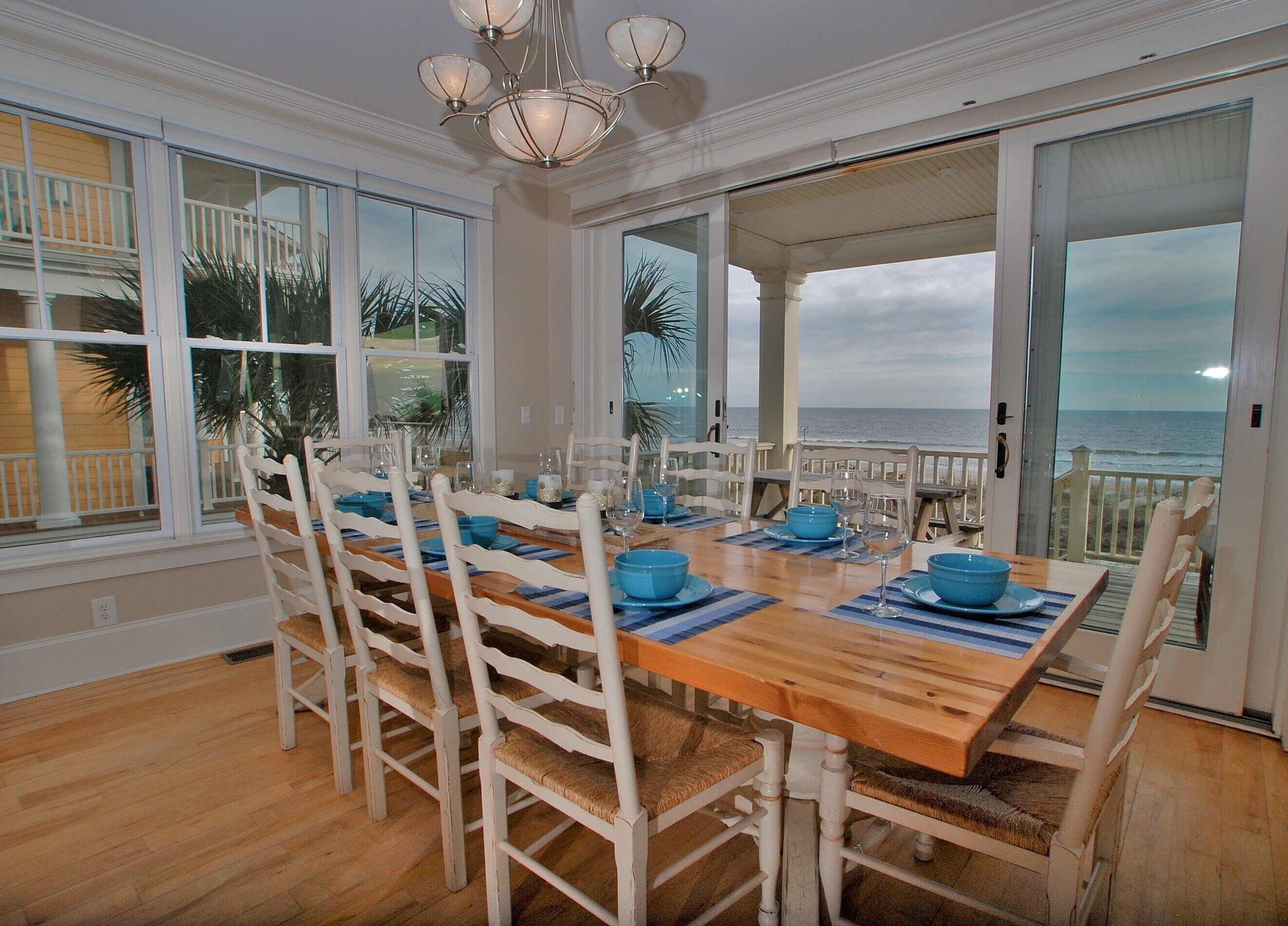 A dining area with a wooden table and white chairs, large windows with a view of the ocean, and a chandelier.