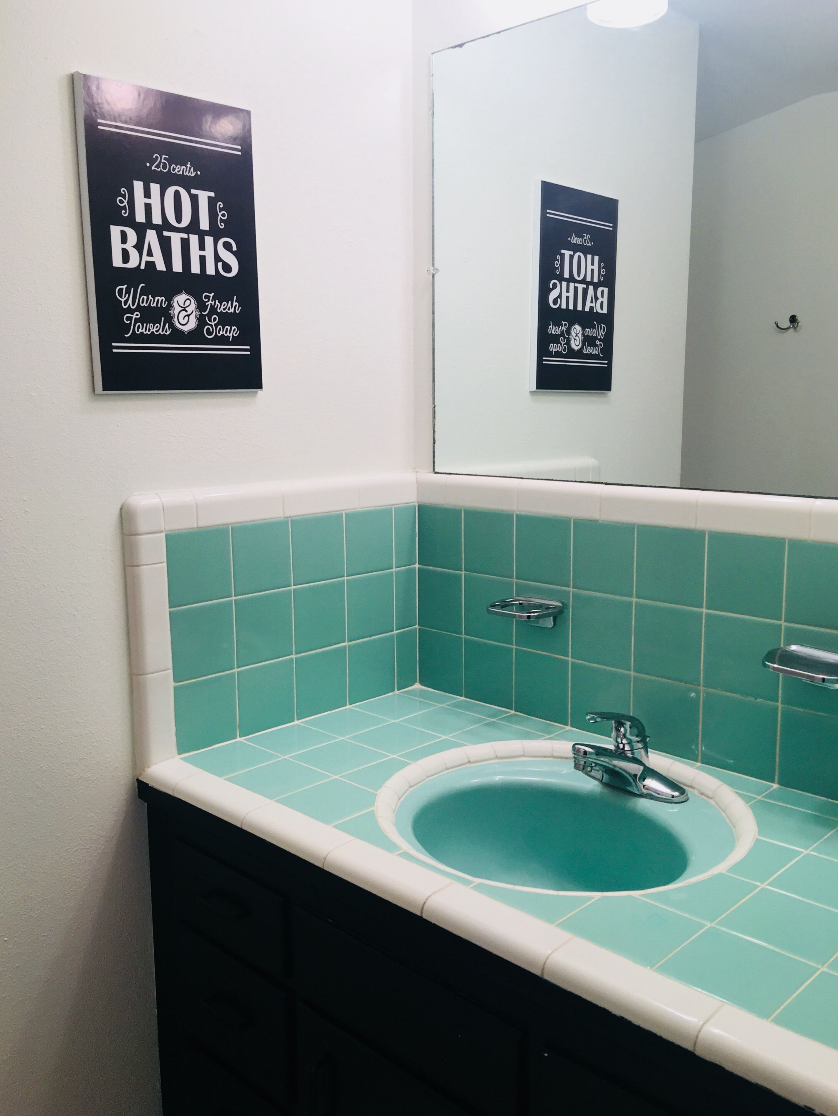 A bathroom with a turquoise sink, white and turquoise tiles, and two signs on the wall advertising 'HOT BATHS' and '25 cents'.