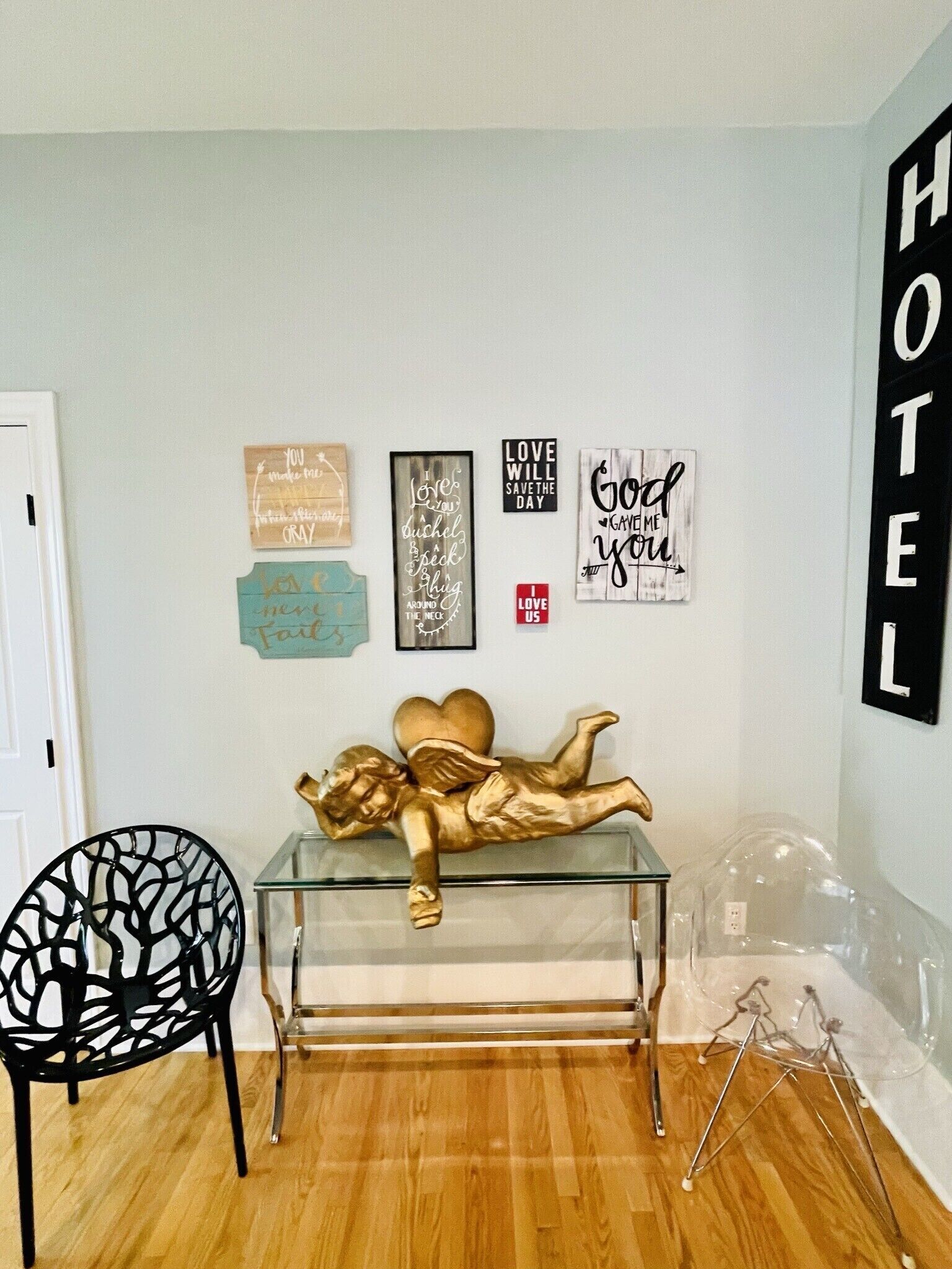 A hotel room with a wooden floor, a black chair, a glass coffee table with a golden angel sculpture, and several wall decorations including motivational quotes and a 'HOTEL' sign.