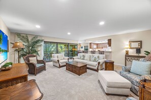 A spacious living room with a large window, a sofa set, a coffee table, and a dining area with wooden cabinets.