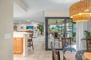 A modern kitchen with a central island, a dining area with wicker chairs, and a view of a tropical landscape through large sliding doors.