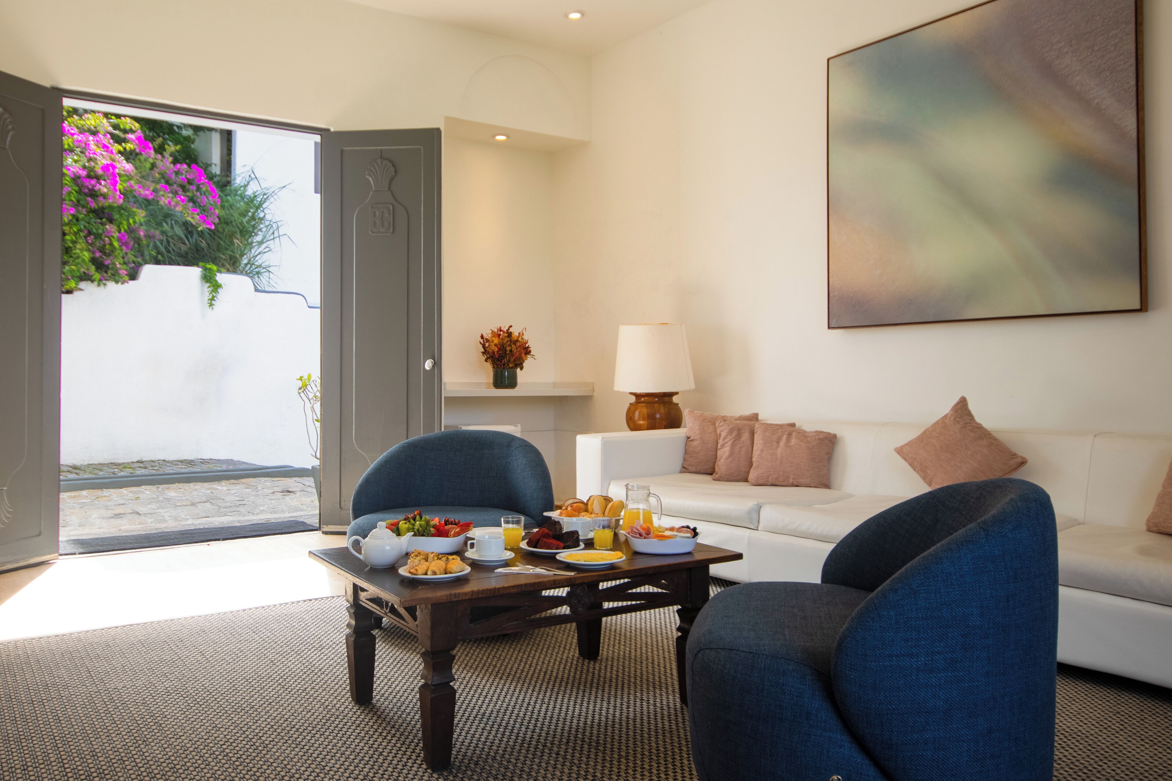 A modern living room with a white sofa, two blue armchairs, a coffee table with breakfast items, and a large abstract painting on the wall.