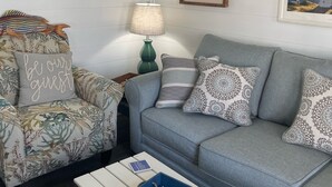 Ein Wohnzimmer mit grauem Sofa, einem floralen Sessel, einem weißen Couchtisch mit Meeressternen und einer Lampe.