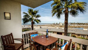 Un balcon avec une table et des chaises en bois, donnant sur une plage bordée de palmiers et offrant une vue sur un ciel dégagé.