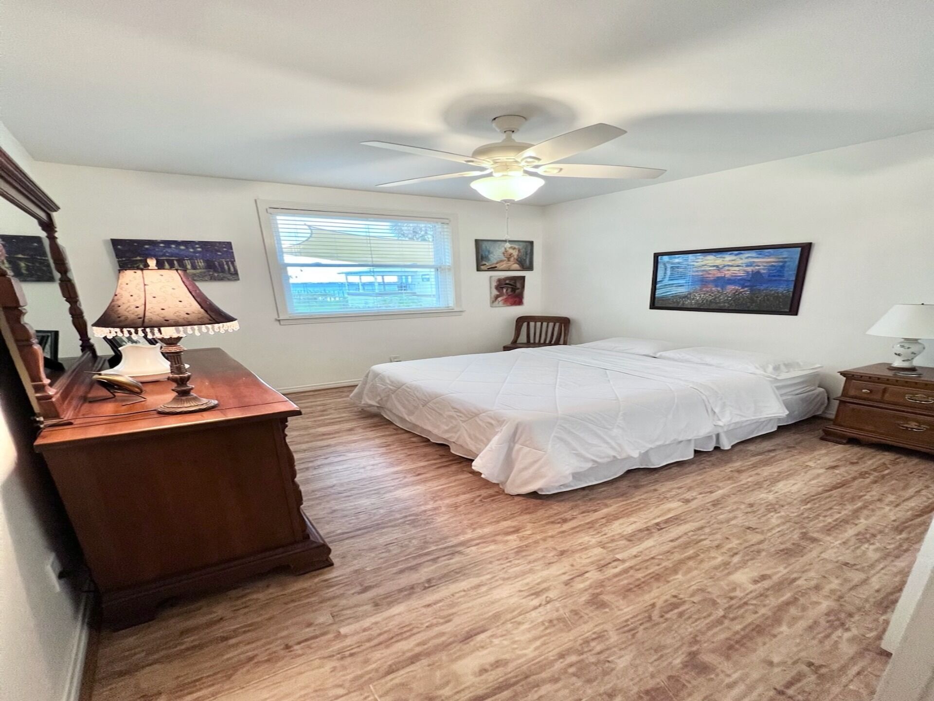 A bedroom with a bed, a ceiling fan, a window with blinds, a wooden dresser, and a lamp.