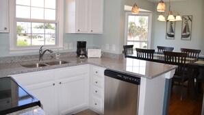 A kitchen with white cabinets, granite countertops, a dishwasher, and a coffee maker. There are two windows providing natural light and a view of the outside. - Oceanfront Cottage with Private Beach Access (Emerald Isle)