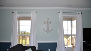 A room with a large window, a black sofa, and a wooden anchor decoration on the wall. - Oceanfront Cottage with Private Beach Access (Emerald Isle)