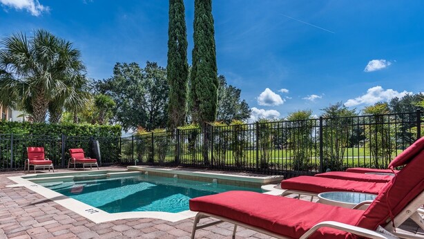 A swimming pool with lounge chairs, a brick patio, and a view of tall trees and a clear sky.