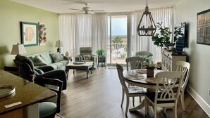 A spacious living room with a green couch, a dining table with chairs, a ceiling fan, and a view of the outdoors through large windows. -  Gorgeous Sunsets!!  We now have some openings for first part of Aug.    (Orange Beach)