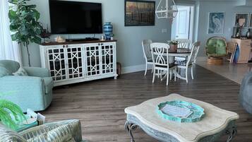 A living room with a TV, a dining area with chairs, a large plant, and a decorative table.