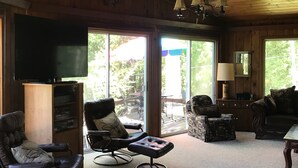 A cozy living room with wooden walls and ceiling, a ceiling fan, and large windows offering a view of the outdoors.