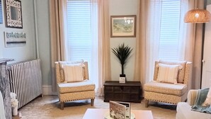 Un salón acogedor con dos sillones, una mesita de centro y una araña. Hay dos ventanas con cortinas, un radiador y un cuadro en la pared.