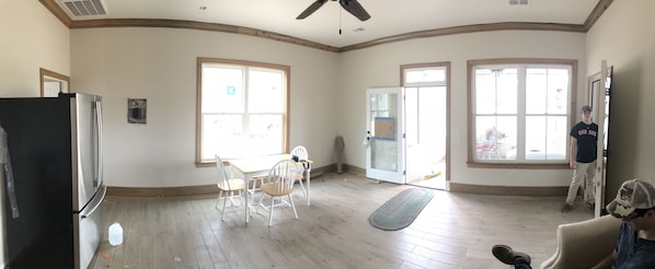 A spacious living room with a large window, a dining area with a table and chairs, a refrigerator, and a ceiling fan.