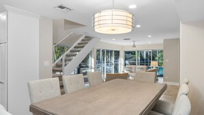 Un espace repas moderne avec une grande table en bois et des chaises rembourrées, un escalier et de grandes fenêtres donnant sur l’extérieur.