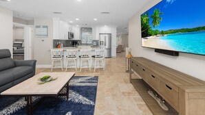 Un salon moderne avec un téléviseur à écran plat, un canapé gris, une table basse et une cuisine en arrière-plan.