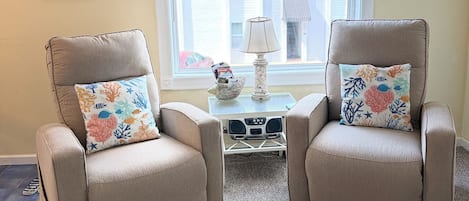 A room with two beige armchairs, a small table with a lamp, and a window with a view of buildings.
