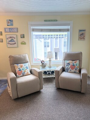 A room with two beige armchairs, a small table with a lamp, and a window with a view of buildings.