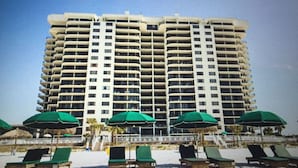 Un hotel moderno de gran altura con varios balcones, sombrillas de playa y sillones de descanso en la planta baja.