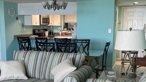 Una sala de estar con sofás a rayas, una mesa de comedor y sillas. Se observa una cocina al fondo con un microondas y un lavaplatos.