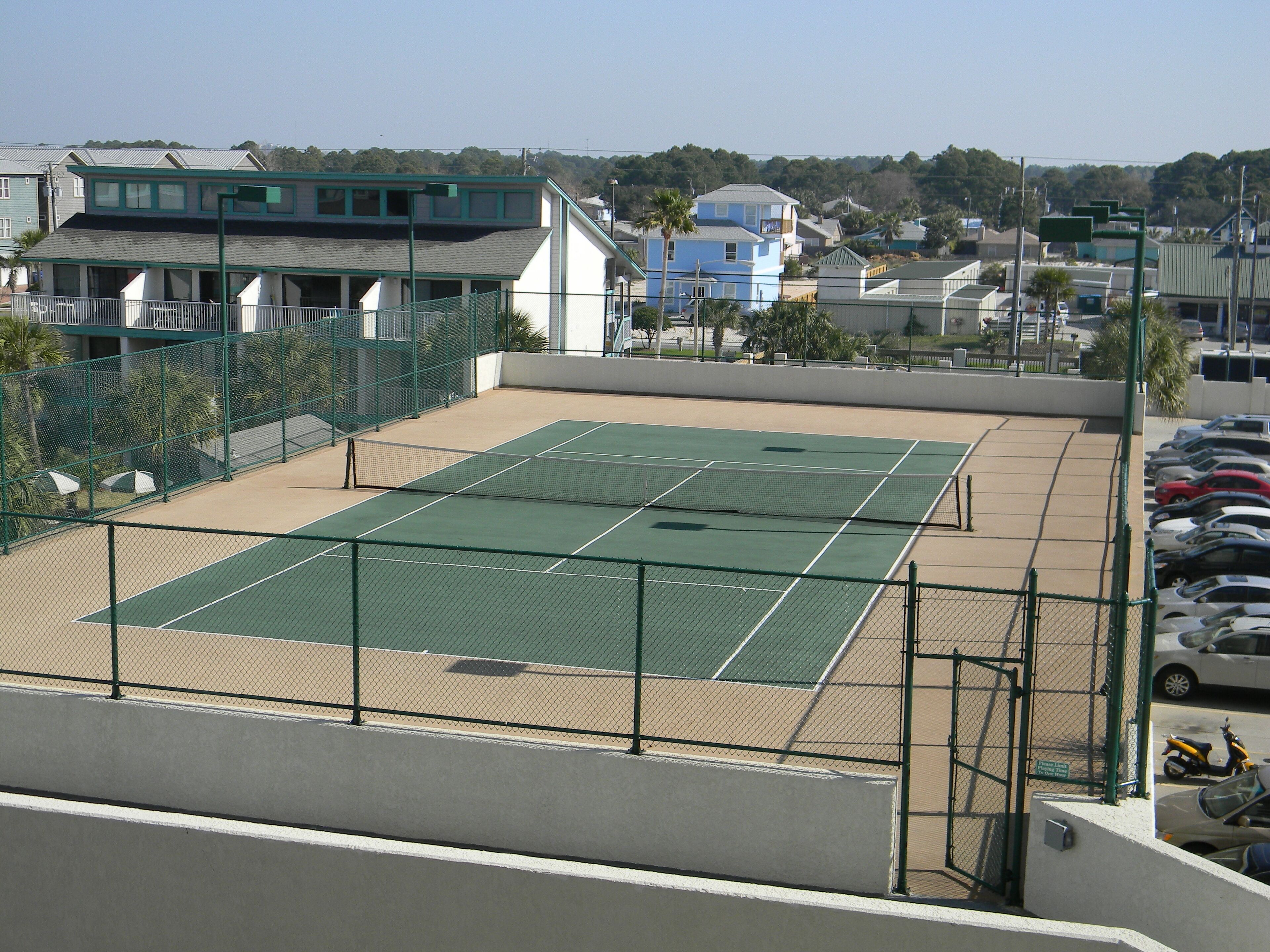 Una cancha de tenis con red, rodeada por una valla, y contigua a un estacionamiento con coches.