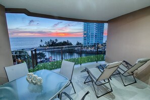 A balcony with a glass table and chairs, overlooking a sunset view of the ocean and a tall building.