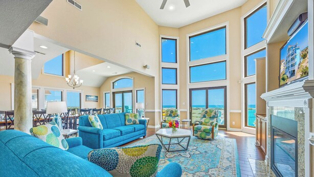 A spacious living room with a large blue sofa, a fireplace, and large windows offering a view of the ocean. - Premier Oceanfront Dog Friendly with Pool, Hot Tub, Elevator, Theater, Tiki Bar (Salvo)