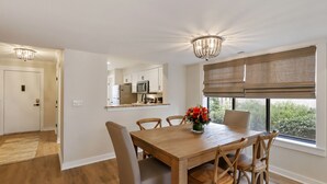 A dining area with a wooden table and chairs, a vase of flowers, and a view of the kitchen through a glass partition. - Courtside 61- Updated Flat - Pool View - Free Tennis (Hilton Head Island)