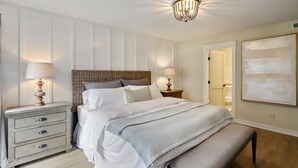 A bedroom with a large bed, two bedside tables with lamps, a bench, and a wall-mounted artwork. - Courtside 61- Updated Flat - Pool View - Free Tennis (Hilton Head Island)