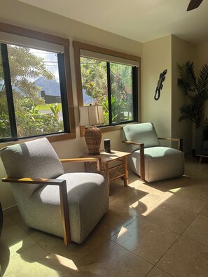 A room with two armchairs, a small table, and a lamp. Large windows provide a view of greenery outside.