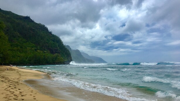 A beach with footprints leading to the water, a forested cliff, and waves crashing onto the shore. - Spacious remodeled town home, beach access,quiet prime location in Princeville (princeville)