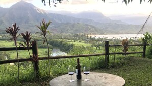 A table with a bottle and two glasses of wine, set against a scenic view of a river and mountains. - Spacious remodeled town home, beach access,quiet prime location in Princeville (princeville)