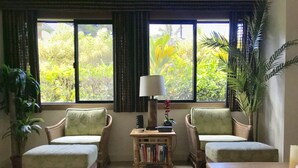 A room with two wicker armchairs, a wooden coffee table, and a small side table with books and a lamp. Large windows with curtains provide a view of greenery outside. - Spacious remodeled town home, beach access,quiet prime location in Princeville (princeville)