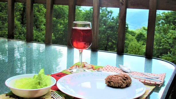 A table with a glass of red wine, a plate with a cookie, a bowl of salad, and cutlery on a glass table with a view of trees and mountains. - ALPINE LOG CABIN/COZY, COMFORTABLE, MODERN/BLUE RIDGE MOUNTAINS NEAR BOONE N.C. (Boone)