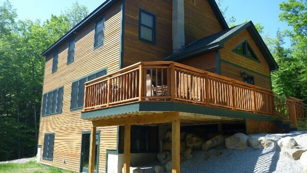 Une cabane en bois à deux étages, dotée d’un balcon, de fondations en pierre et entourée d’une zone recouverte de gravier.