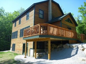 A two-story wooden cabin with a balcony, a stone foundation, and a surrounding gravel area. - Privacy, Spacious Home, Minutes to Attitash, Rocky Branch Trail, Storyland. (Bartlett)