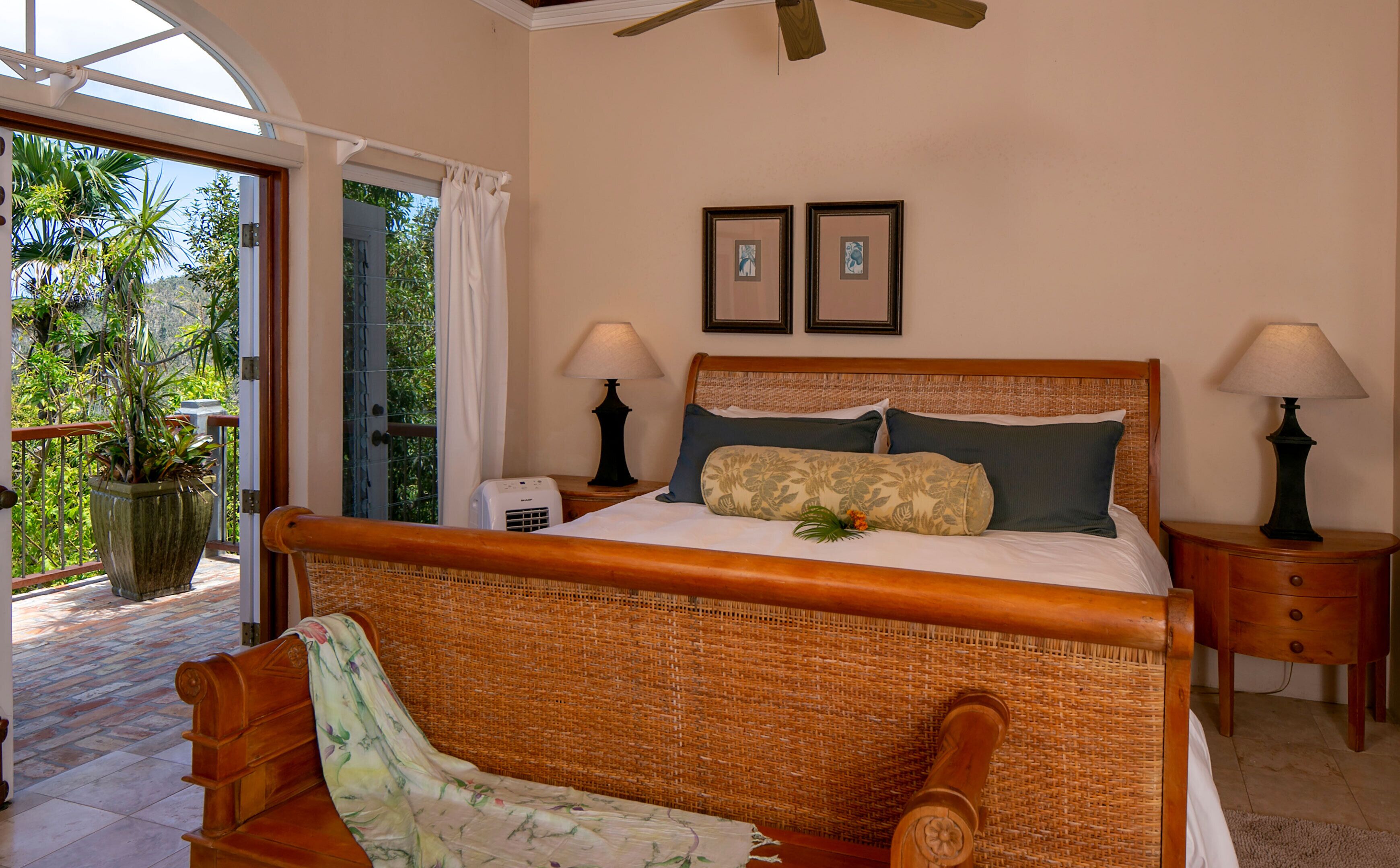 A room with a bed, two bedside lamps, a potted plant, and a view of the outdoors.