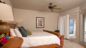 Une chambre à coucher avec un lit en bois, un ventilateur de plafond, une œuvre d’art encadrée et une vue à travers une porte vitrée.